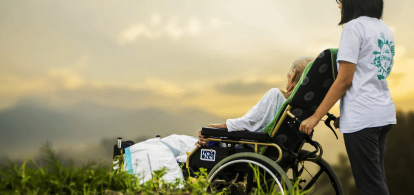 A bedsores patient sitting on a wheelchair with a nurse behind taking a view outside.
