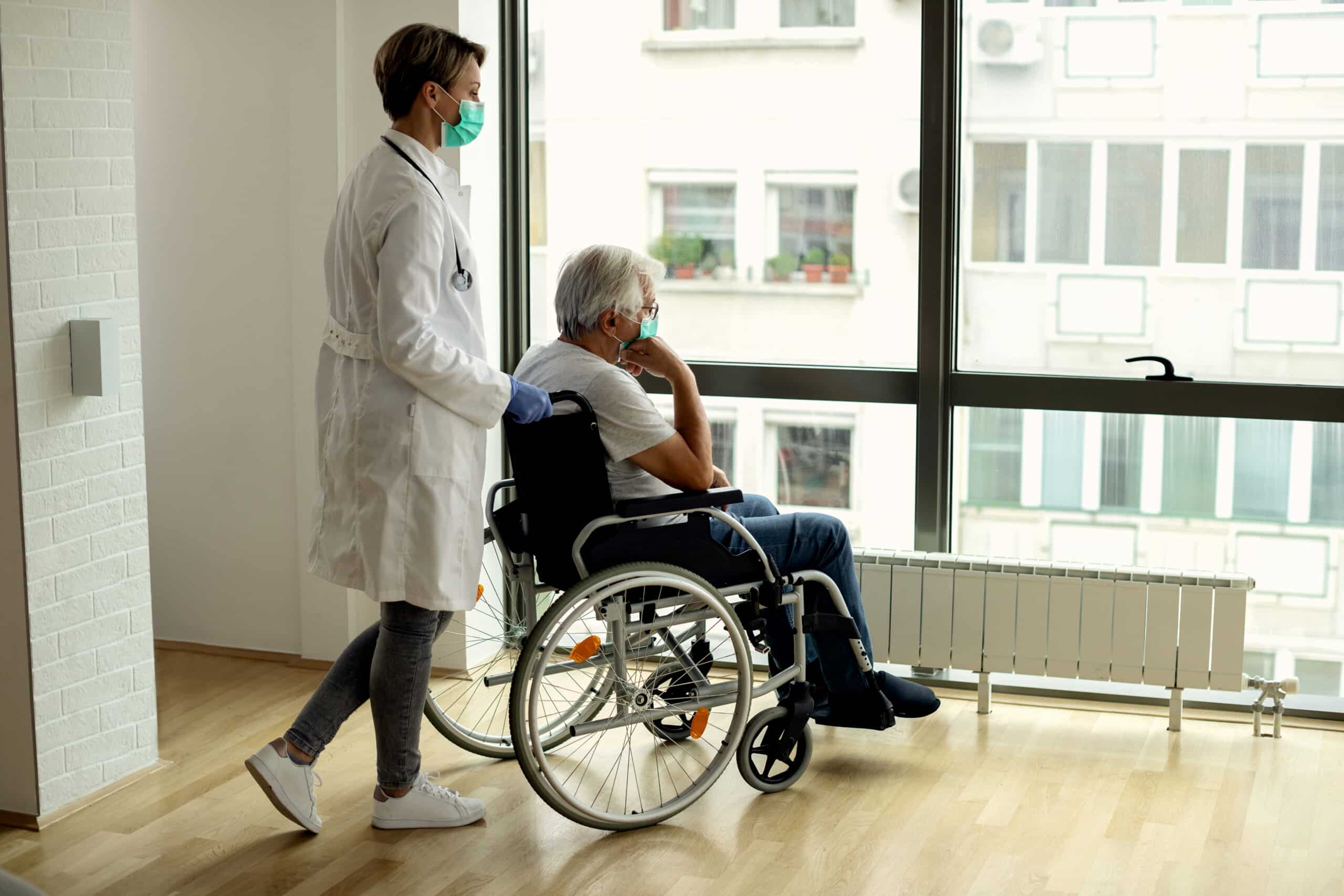 Female doctor pushing senior man in wheelchair to the window. They are wearing protective face masks.