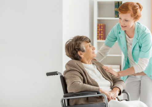 A nurse smiling with an elderly woman that is sitting on a wheelchair in a nursing home.